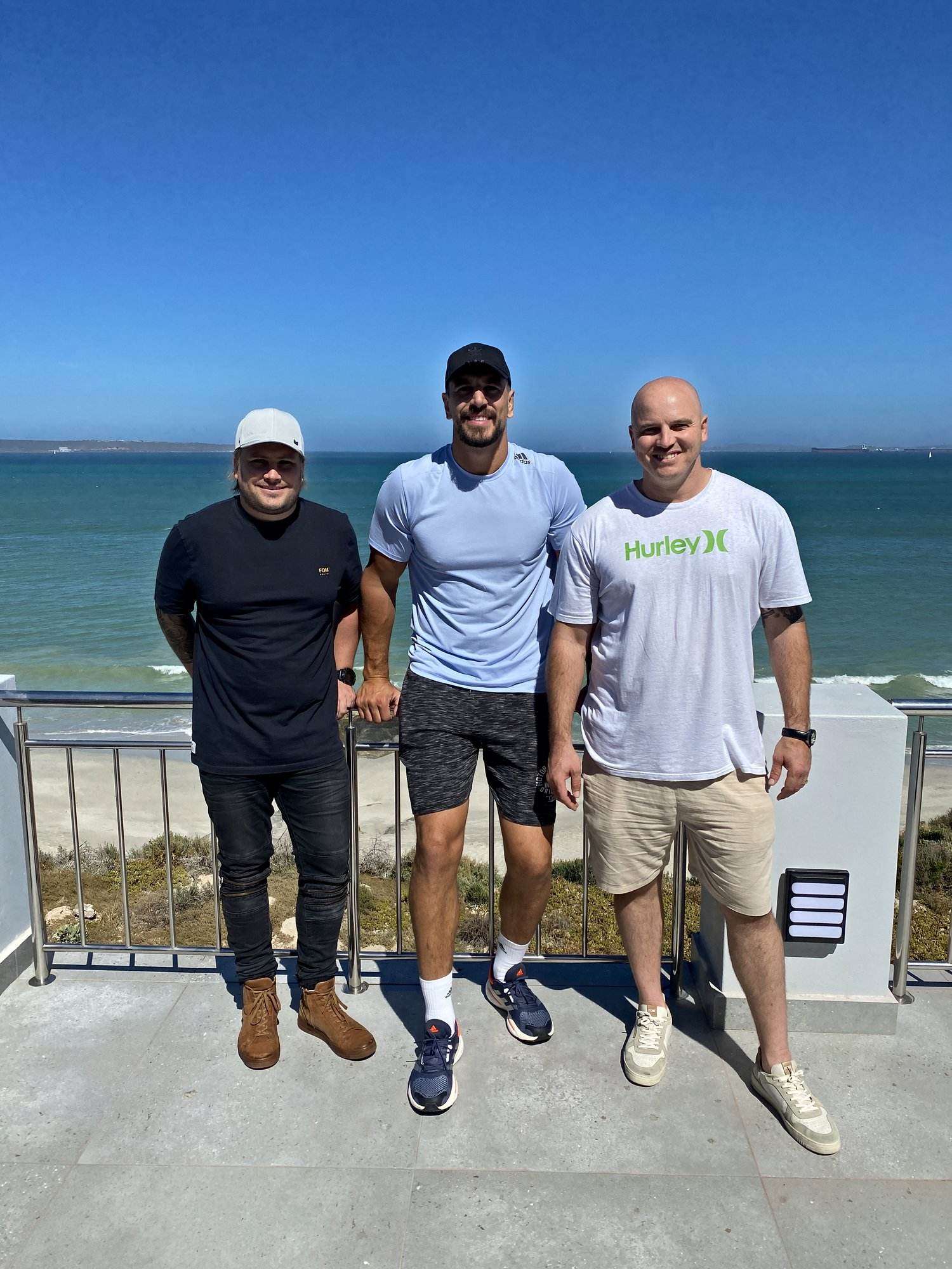 Eben Etzebeth × Made Dot campaign: three men standing on a sunny balcony with the ocean behind them in Langebaan