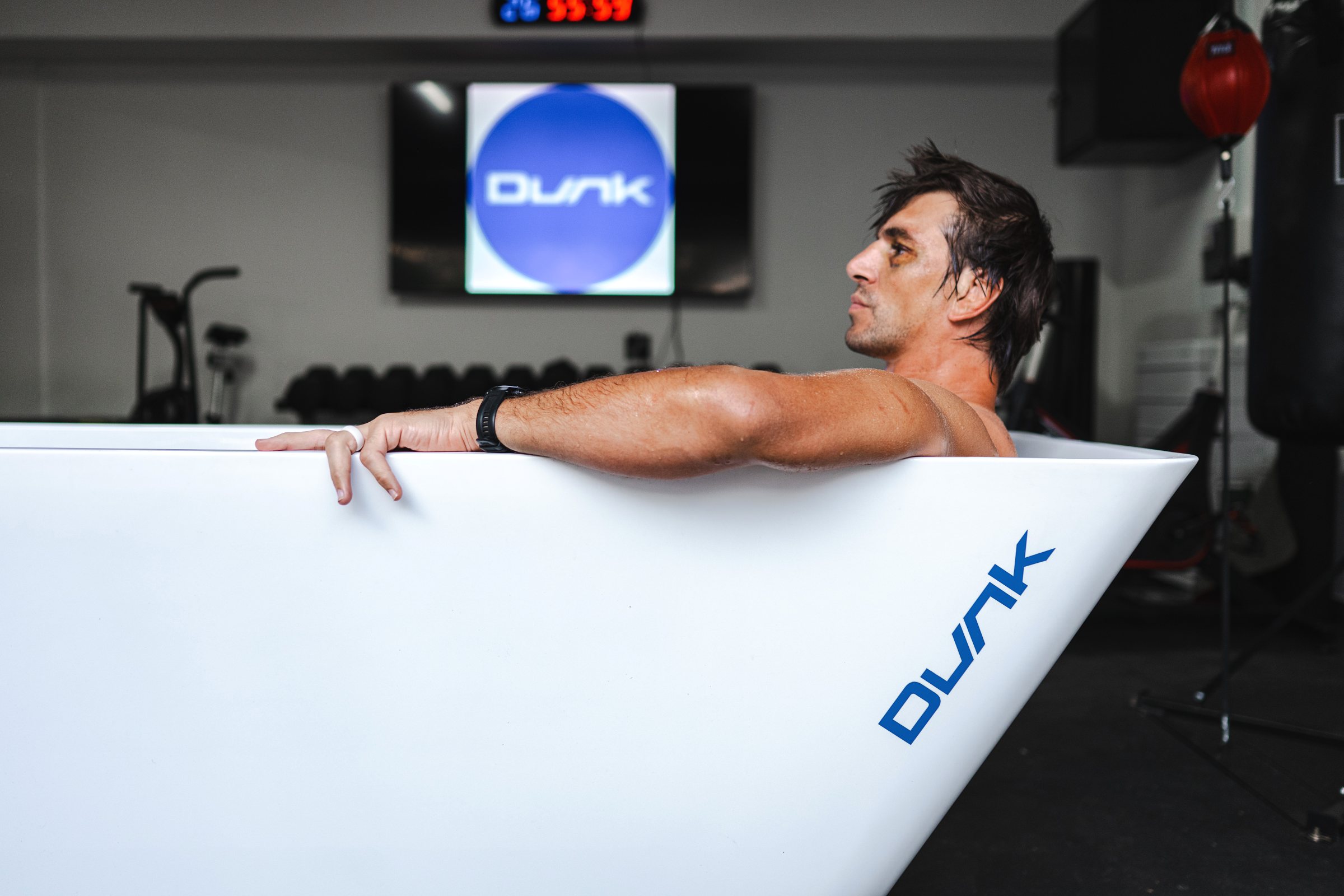 Man relaxing in a white Dunk ice bath in a home gym, with the Dunk logo on screen behind