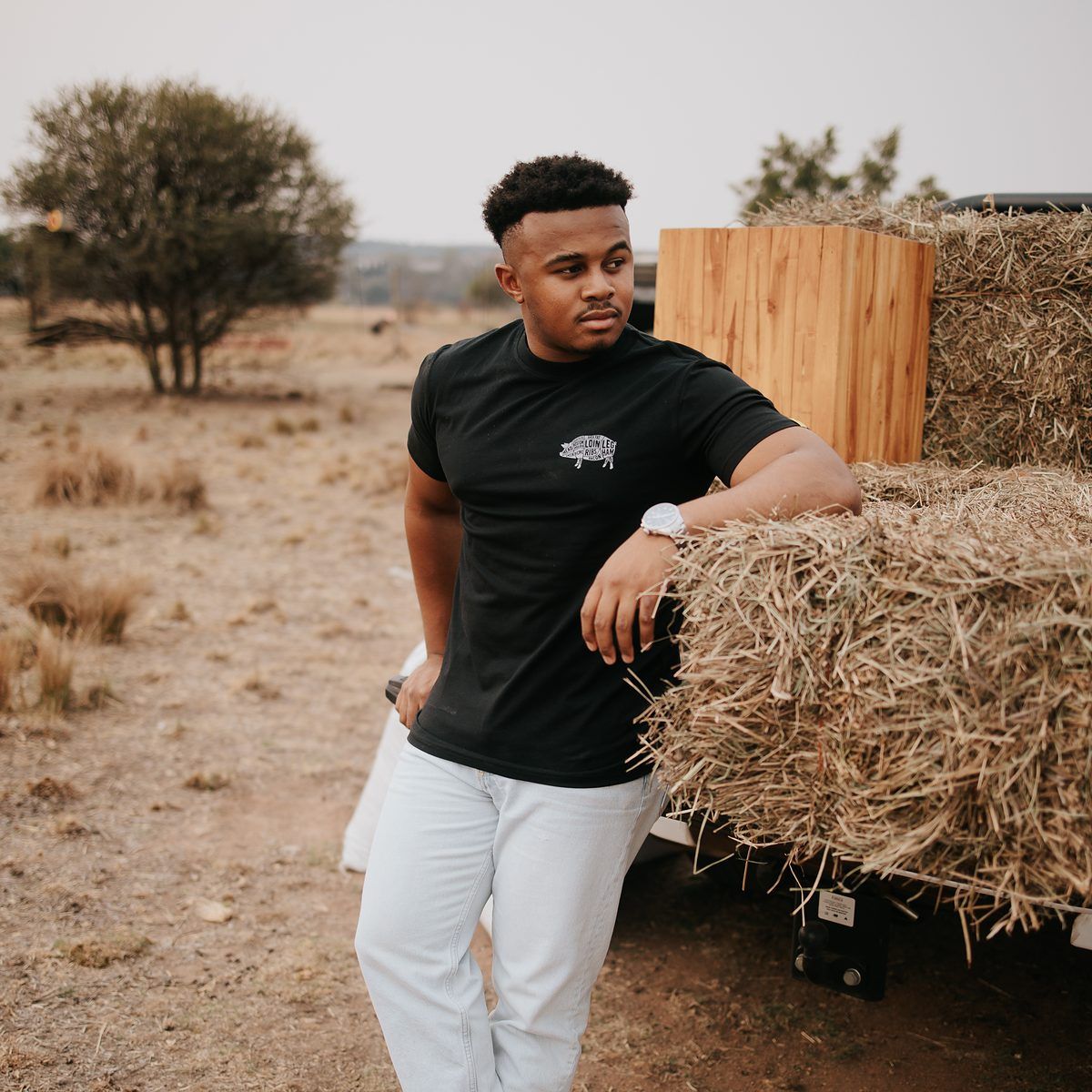 Mieliestronk apparel brand shoot: model in a black Mieliestronk tee leaning on hay bales on a farm